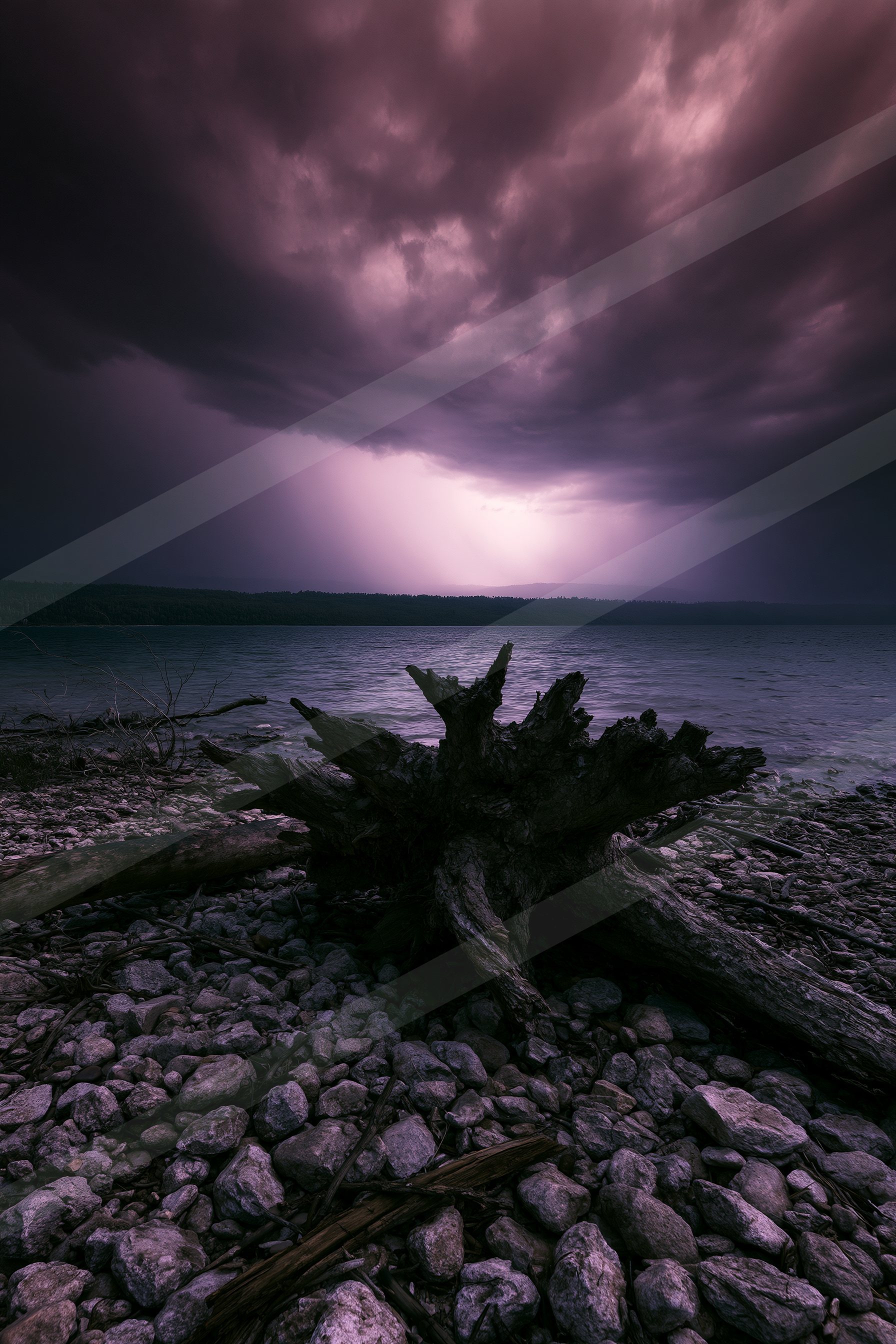 A serene lake under a dark, stormy sky with lightning illuminating the horizon. A weathered log lies prominently on a rocky shoreline.