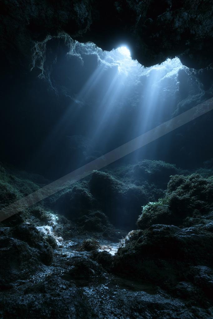Strong beams of sunlight streaming through an opening at the top of a dark, rocky cave onto a wet, moss-covered floor.
