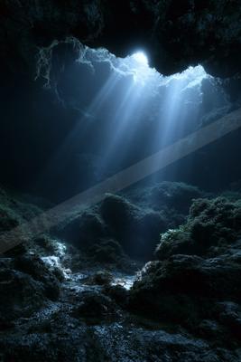 Strong beams of sunlight streaming through an opening at the top of a dark, rocky cave onto a wet, moss-covered floor.