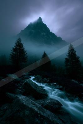 A dramatic mountain peak shrouded in thick fog rises above a flowing stream that cascades over rocks in the foreground, surrounded by dark evergreen trees in a 