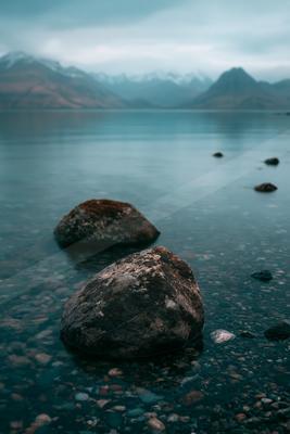 A serene lake scene featuring moss-covered rocks on a pebbled shore, with distant snow-capped mountains under an overcast sky.