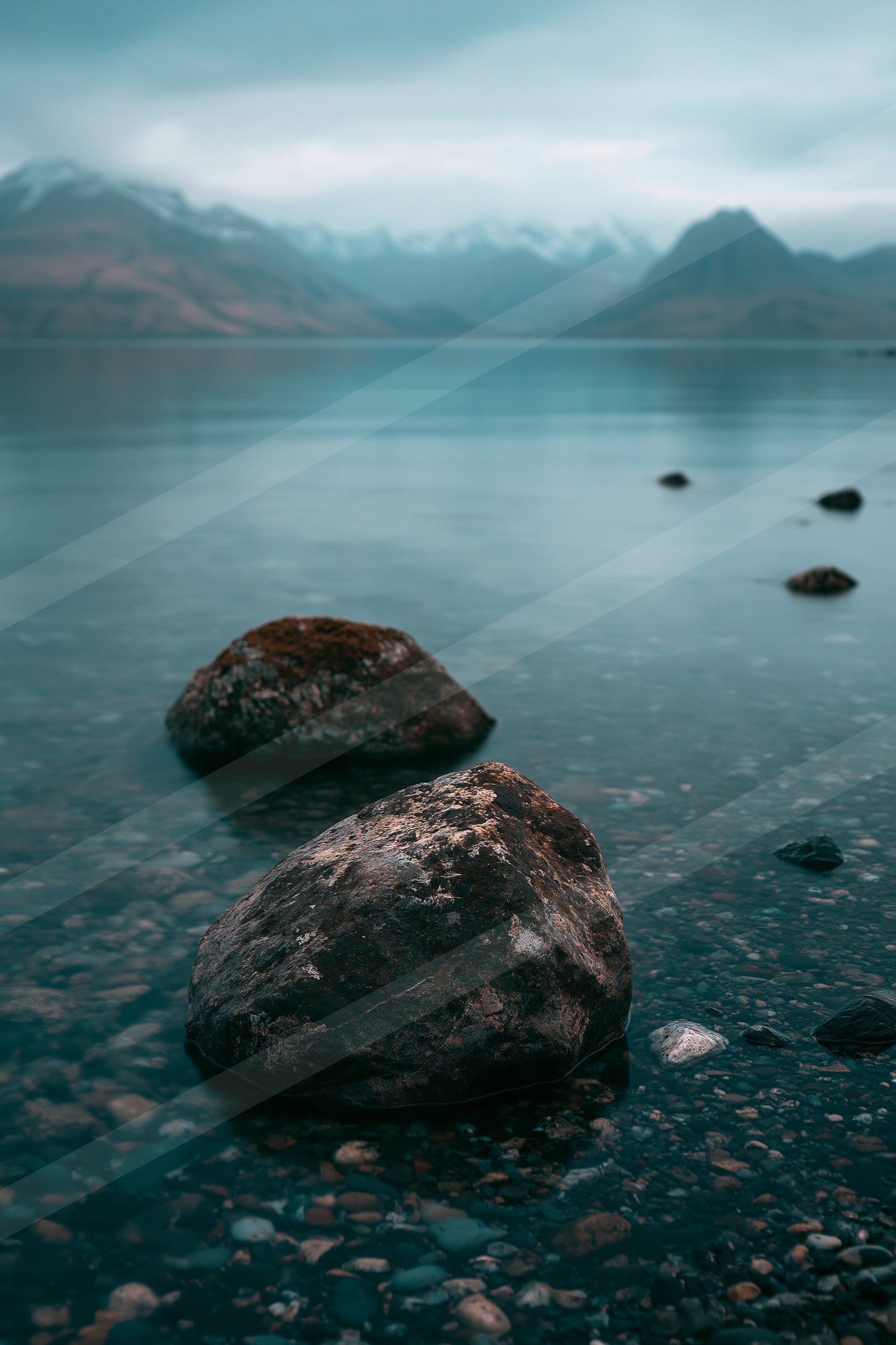 A serene lake scene featuring moss-covered rocks on a pebbled shore, with distant snow-capped mountains under an overcast sky.