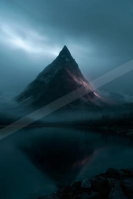 A sharp, rocky mountain peak rises through thick fog above a calm alpine lake, its reflection mirrored perfectly in the still water under a moody blue sky.