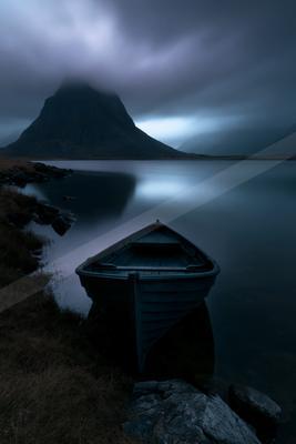 A small blue rowboat rests on the shore of a calm lake with a large misty mountain rising in the background under a dark, overcast sky.