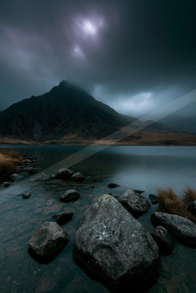A dramatic landscape featuring a calm lake with large rocks in the foreground, a rugged mountain partially shrouded in mist, and a dark, stormy sky with a brigh