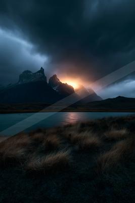 A jagged mountain peak illuminated by a fiery sunset glow piercing through dark storm clouds, reflected in a calm lake surrounded by dry grass in the foreground