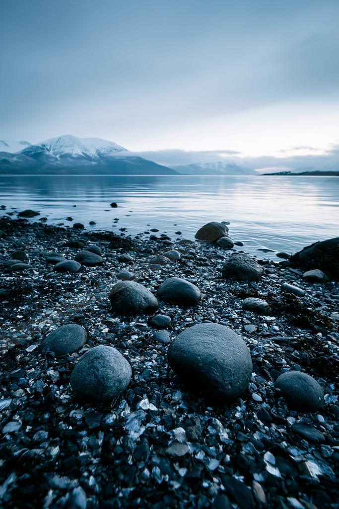 A pebble and rock strewn lakeshore in the foreground with calm water stretching to snow-capped mountains under a muted blue sky.