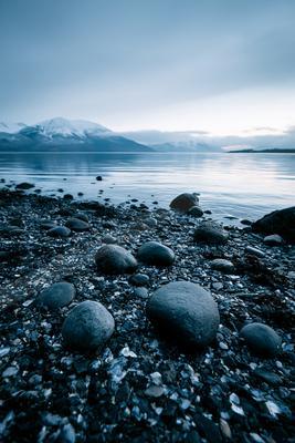 A pebble and rock strewn lakeshore in the foreground with calm water stretching to snow-capped mountains under a muted blue sky.