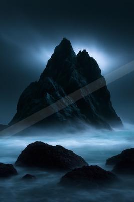 A tall, jagged rocky sea stack rises from a misty ocean under a dark, moody sky, with foreground rocks and smooth, ethereal water created by long exposure.