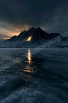 Dramatic snowy mountain range with sunlight breaking through a gap in the peaks, casting a golden reflection on the surface of a vast frozen lake under a moody 