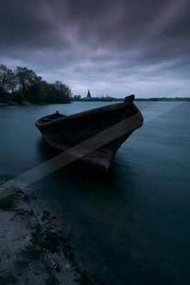 An old weathered wooden boat rests partially on a sandy shore in dark, still water under a moody, overcast sky with a distant shoreline and structures visible.