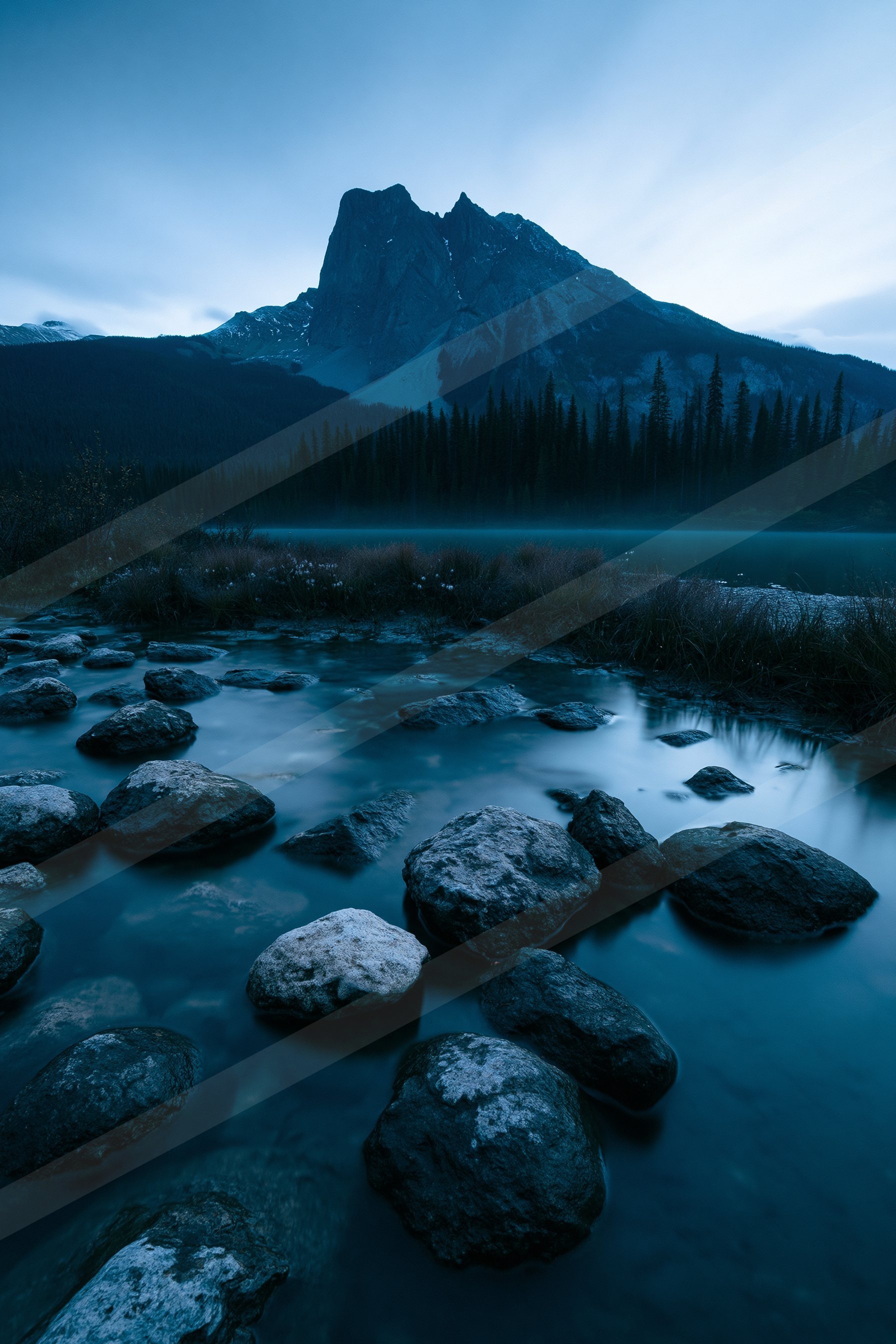 A tranquil lake surrounded by rocks, evergreen trees, and a snow-capped mountain under a dusky sky.