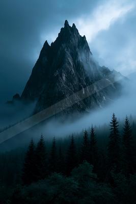 A jagged rocky mountain peak shrouded in thick blue mist and low clouds, towering over a dense forest of dark evergreen trees in the foreground.