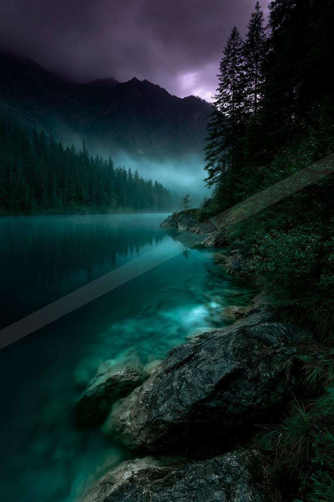 A serene turquoise lake reflects a dense coniferous forest and rugged mountains under a dramatic purple and dark cloudy sky, with large rocks in the foreground
