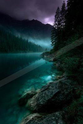 A serene turquoise lake reflects a dense coniferous forest and rugged mountains under a dramatic purple and dark cloudy sky, with large rocks in the foreground 