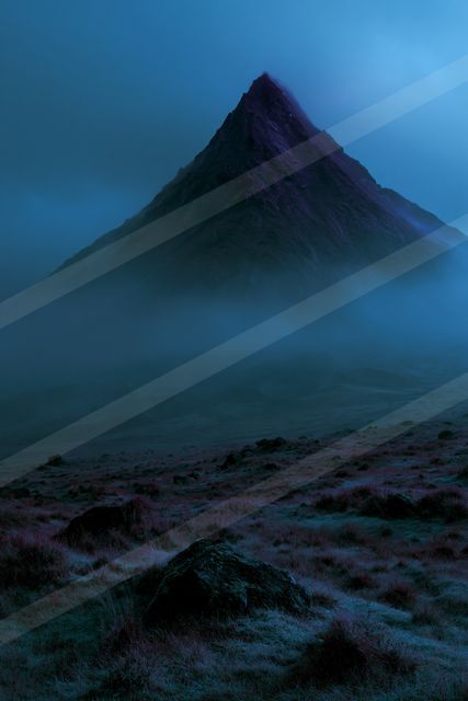 A towering mountain peak rises sharply against a twilight sky, partially obscured by swirling mist. The foreground features rocky terrain under a moody blue-pur