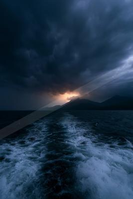A turbulent dark blue sea with a foamy wake in the foreground under a sky filled with heavy storm clouds, with a break of warm light on the horizon behind silho
