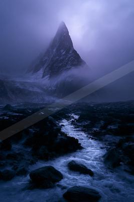 A sharp rocky mountain peak rises through thick purple mist above a turbulent stream flowing through a boulder-strewn foreground.