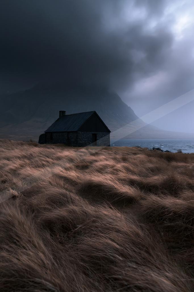 A dark stone cottage stands in a field of tall brown grass beside a lake, with a large mountain looming under heavy storm clouds.