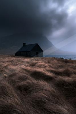 A dark stone cottage stands in a field of tall brown grass beside a lake, with a large mountain looming under heavy storm clouds.