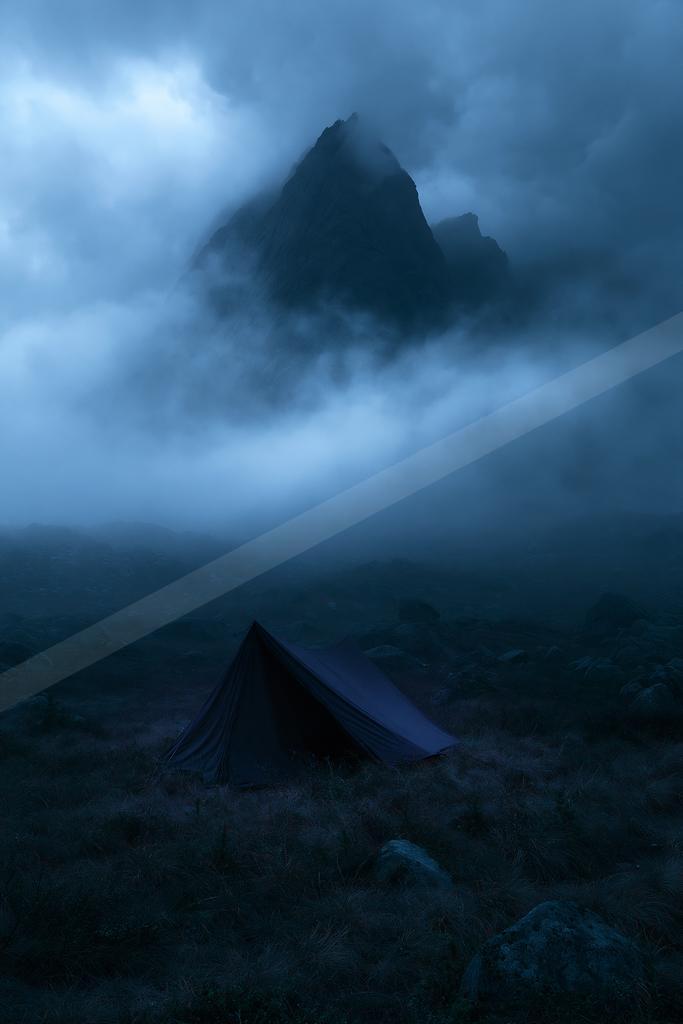A dark camping tent stands in a rocky, grassy foreground with thick fog swirling around a dramatic mountain peak under a moody blue night sky.