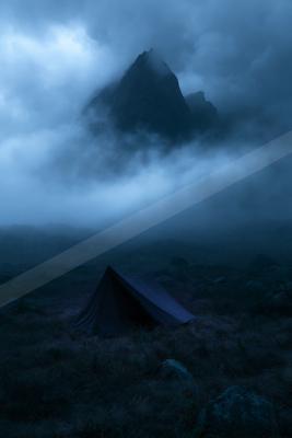 A dark camping tent stands in a rocky, grassy foreground with thick fog swirling around a dramatic mountain peak under a moody blue night sky.