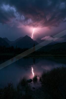 A dramatic lightning bolt strikes a mountain peak during a thunderstorm at night, its bright pinkish light reflecting on the calm surface of a foreground lake s