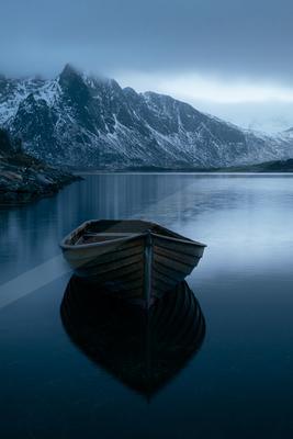 A wooden rowboat floats peacefully on a still lake, surrounded by snow-covered mountains under a clear blue sky.