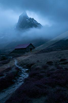 A small stone cabin with a red metal roof sits in a barren, rocky landscape with purple vegetation and a winding path, while a towering snow-capped mountain pea