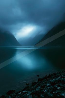A serene lake reflects a bright break in the dark, foggy clouds between steep, forested mountains under a dramatic blue atmosphere.
