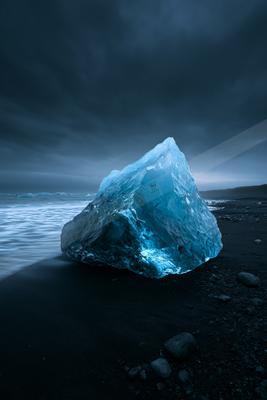 A massive, translucent blue iceberg rests on a dark volcanic sand beach, illuminated against a moody, overcast sky with gentle waves lapping at the shore.