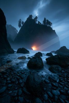 A dramatic night scene of a rocky coastline with large dark boulders in the foreground, misty water flowing around them, and a prominent sea stack covered in tr