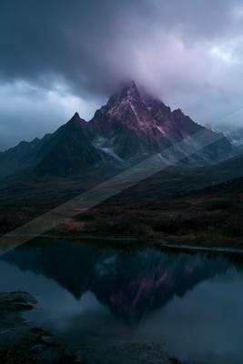 A jagged mountain peak with pink and purple hues rises into thick clouds, perfectly reflected in a calm foreground lake surrounded by dark tundra.