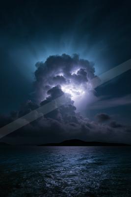 A massive cumulonimbus cloud illuminated by bright lightning strikes over a dark ocean, with a silhouetted landmass on the horizon under a stormy night sky.