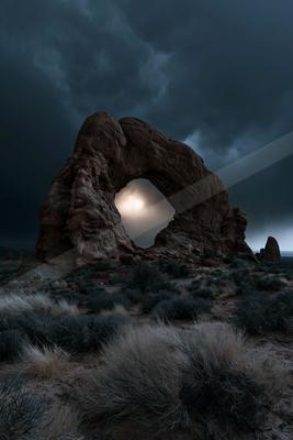 A large sandstone rock arch stands in a desert landscape under dark, stormy clouds, with a bright light visible through its opening.