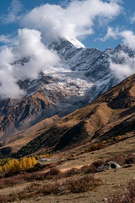 A majestic snow-covered mountain peak partially shrouded in white clouds rises above a vast range of brown and rocky slopes, with an autumn-colored valley featu