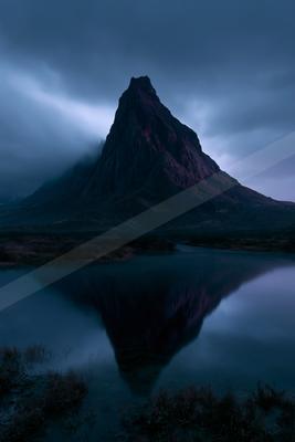 A sharp rocky mountain peak rises above low clouds and is perfectly reflected in the still surface of a foreground lake under a dark, overcast sky.