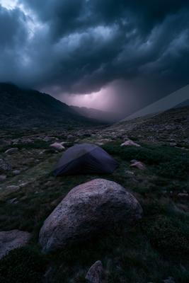 A dark camping tent pitched on a rocky mountainside under heavy storm clouds with lightning visible in the distance.