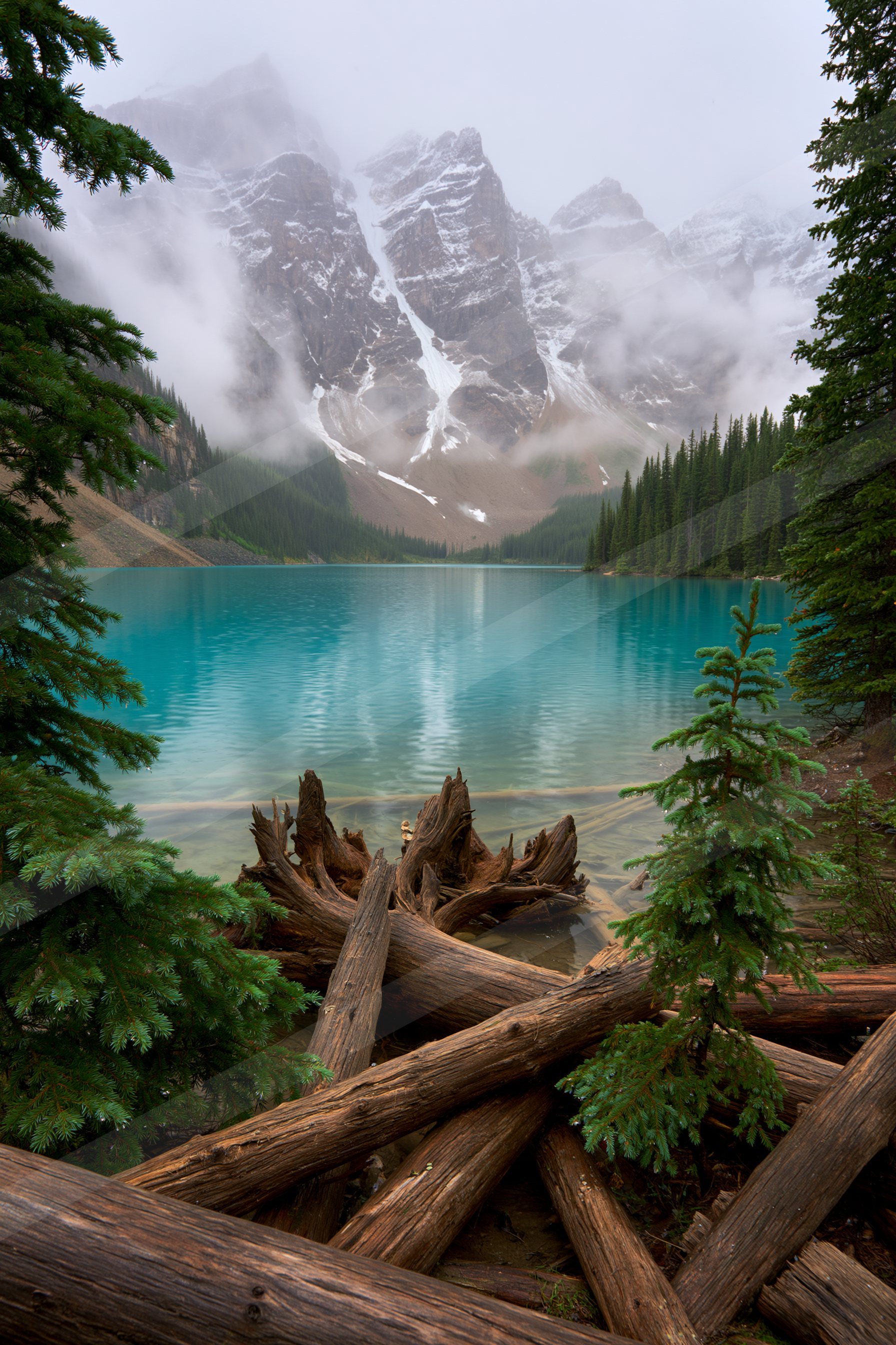 A serene view of a turquoise lake surrounded by evergreen trees, with mist-covered mountain peaks reflected on the water's surface.