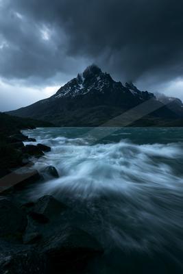 A turbulent turquoise lake with white-capped waves crashes against dark rocks in the foreground, with a jagged snow-dusted mountain peak rising under heavy stor