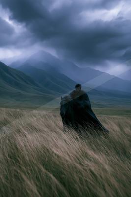 A man wearing a dark fur cloak rides a black horse through tall dry grass in a vast mountainous landscape under a stormy sky.