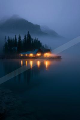 A serene cabin glowing warmly at dusk, reflected perfectly in a still lake surrounded by fog and evergreen trees.