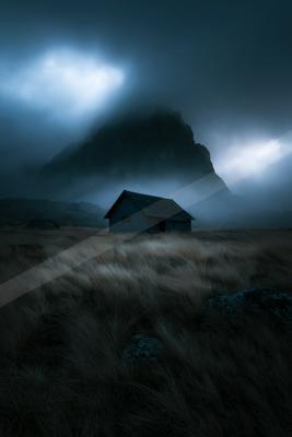 A solitary dark cabin stands in a field of tall dry grass with a large rocky mountain shrouded in thick fog and dramatic clouds in the background.