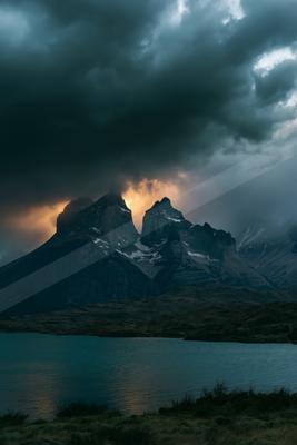 Rugged mountain peaks with patches of snow rise above a calm lake under a dark, brooding stormy sky with orange light breaking through the clouds.