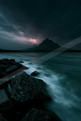 A dramatic mountain peak rises in the distance under a dark, stormy sky with pink and purple hues at dusk. In the foreground, smooth, misty water flows around l