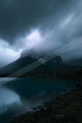 A rugged mountain peak partially shrouded in thick fog rises above a serene turquoise lake under a dramatic, overcast sky, with the mountain's reflection visibl