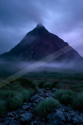 A rugged mountain peak shrouded in thick mist and low clouds under a dark blue twilight sky, with a rocky path and green grass in the foreground.