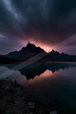 A jagged mountain range is silhouetted against a dramatic twilight sky with pink and purple hues, perfectly reflected in the calm surface of a foreground lake, 