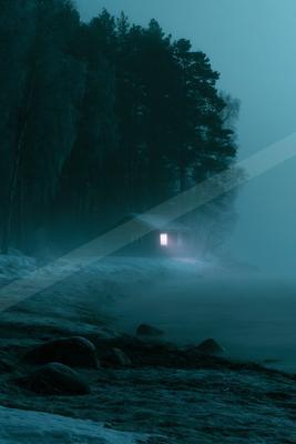 A wooden cabin with a single illuminated window stands on a snowy shore beside a misty lake, surrounded by a dark forest under a blue night sky.
