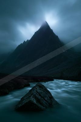 A sharp rocky mountain peak rises through thick fog under a dark cloudy sky, with a smooth flowing river and large foreground rock in a moody blue-toned wildern